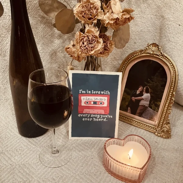 A Valentine themed art print surrounded with a framed photo of a couple, a heart-shaped candle, a wine glass, and dried flowers