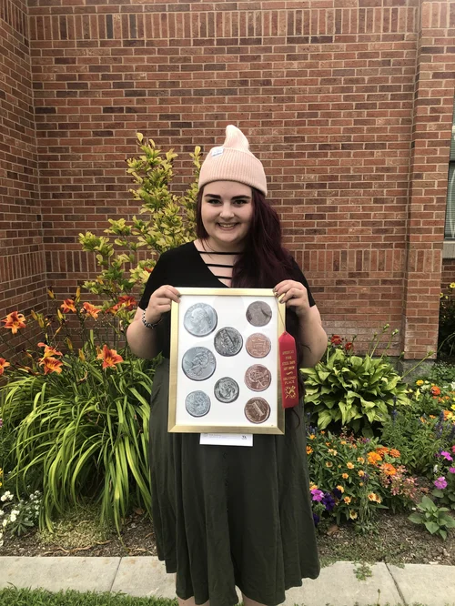 Elise Jackman in front of a building holding an original artwork depicting various coins with an award ribbon attached to the frame