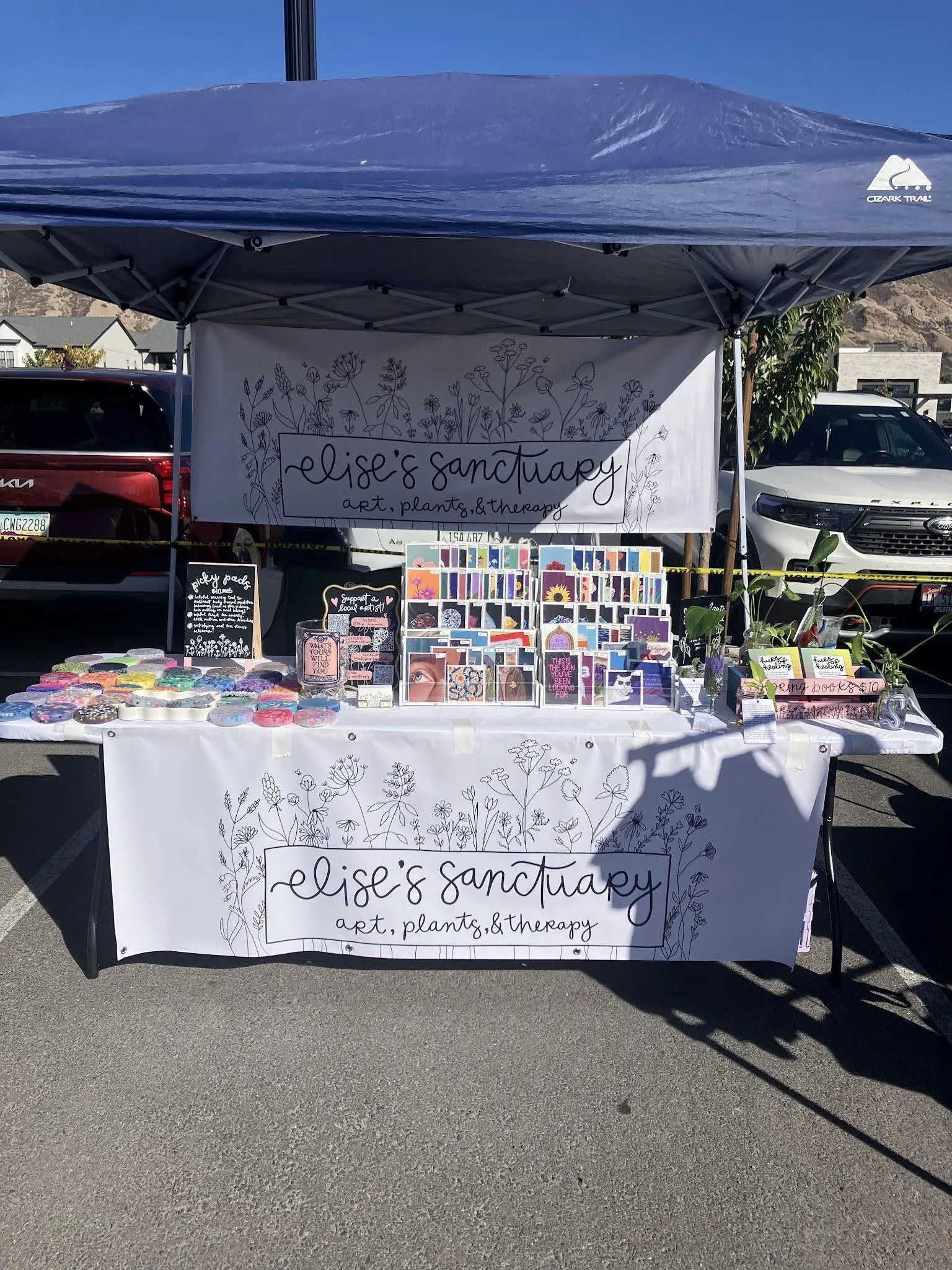 A setup white folding table with several items on it, including handmade picky pads, art prints, and multiple copies of the coloring book Hues with Healing, along with banners both above and below the table, depicting flowers along with the text "Elise's Sanctuary" and "arts, plants, & therapy"