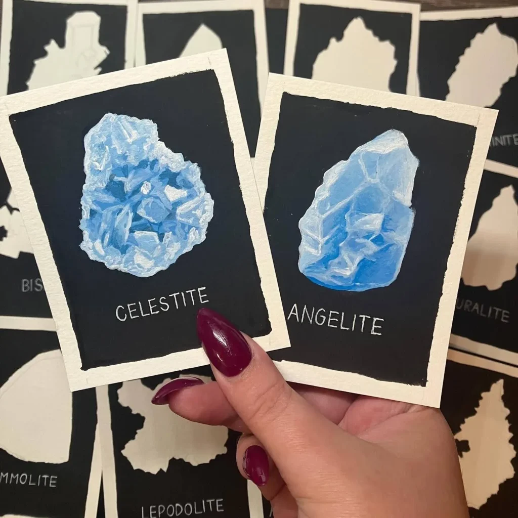 Elise holding original paintings of Celestite and Angelite gemstones, with a backdrop of multiple unfinished gemstone paintings